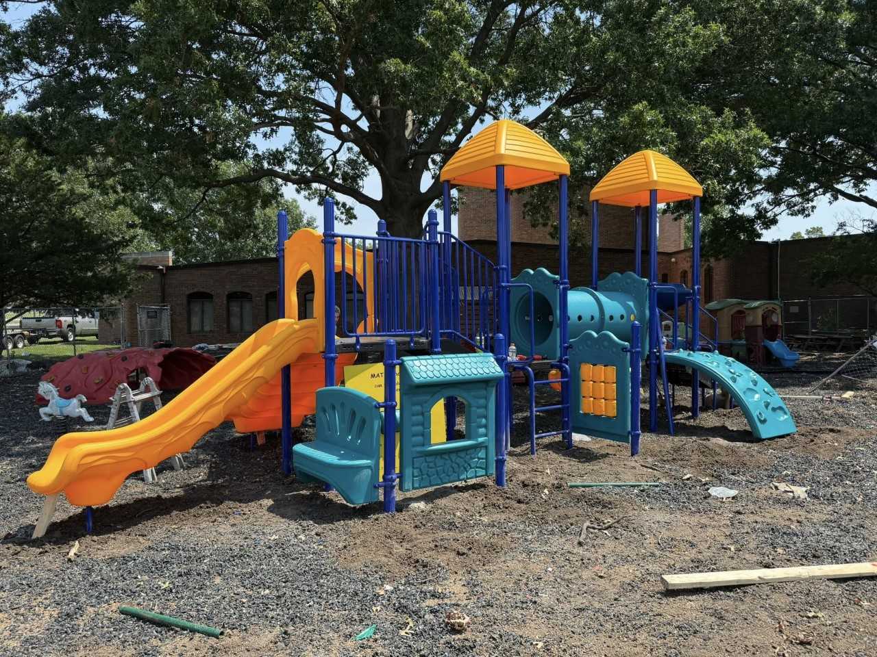 Colorful playground installation with slides, climbing structures, and play panels set outdoors under large trees, designed for children’s active play and exploration.